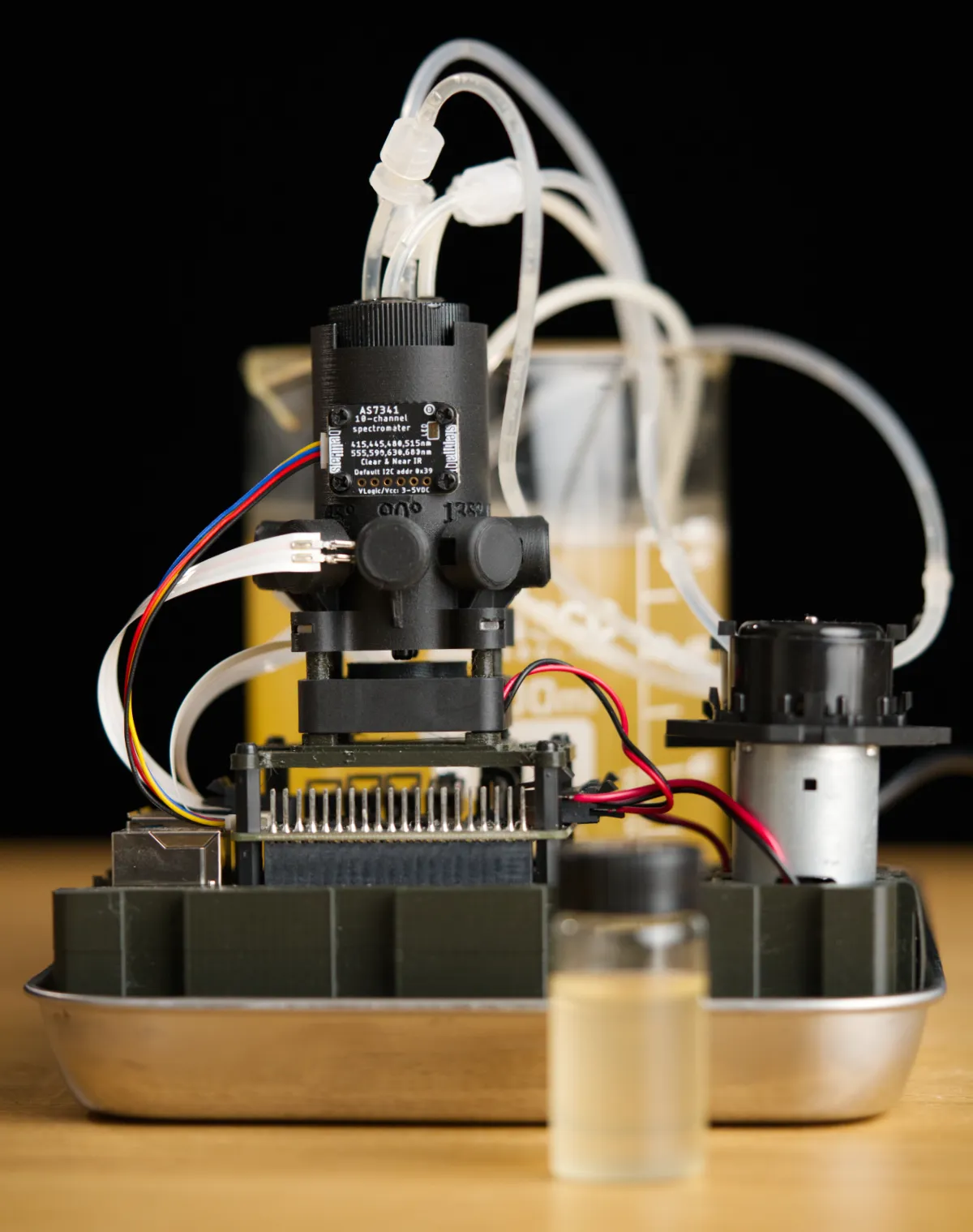 Photograph of pioreactor 20ml v1.0 configured as a flow through cell with a 1L beaker of Kombucha in the background and a 20ml vial of Kombucha in the foreground