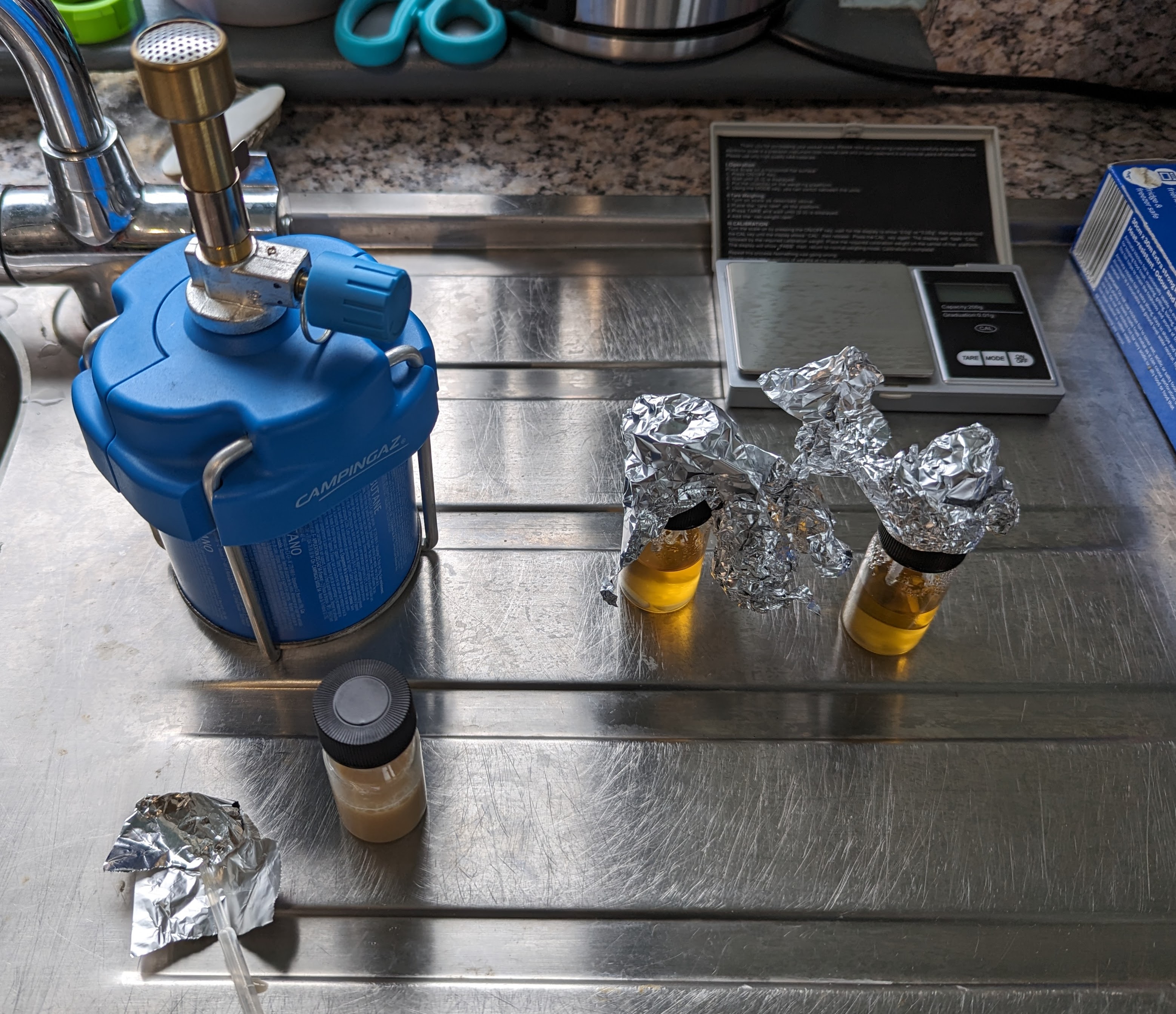 A bunsen burner, small weight scale, three vials and a dropper. Two of the vials contain YPD broth and are covered with aluminium foil. The other vial contains stock solution.